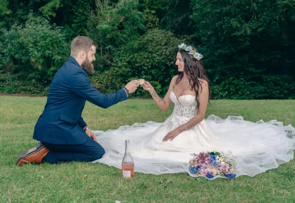 Bride and groom raise a toast on the grass, her skirt spread around her and her bouquet beside them