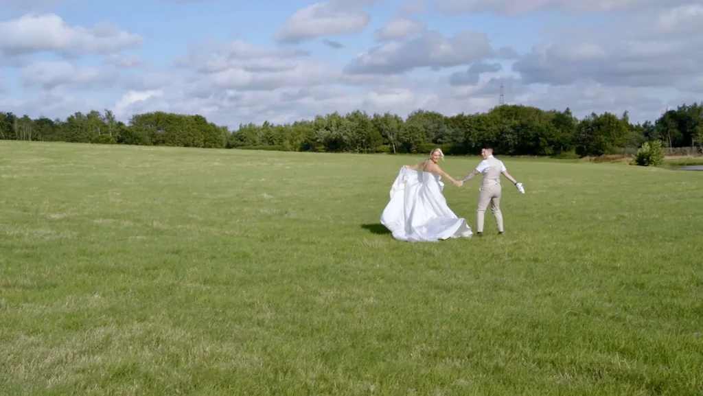 Two brides walk hand in hand across a wide parkland lawn, one in a white gown and one in a beige waistcoat and trousers
