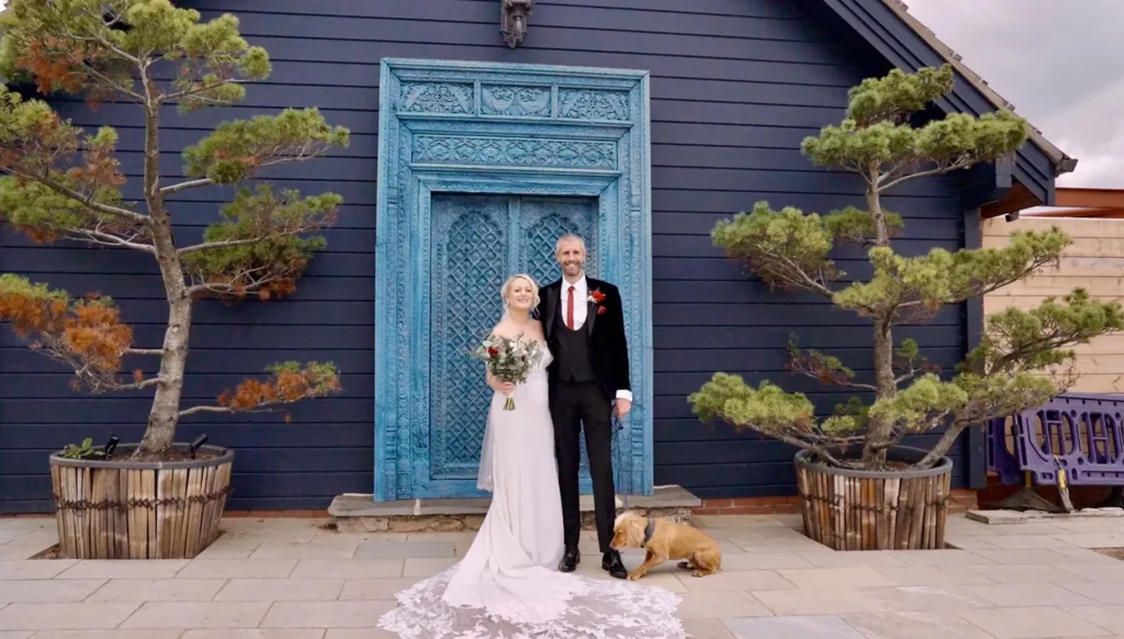 Bride and groom with their dog in front of an ornate blue door, a small pine either side