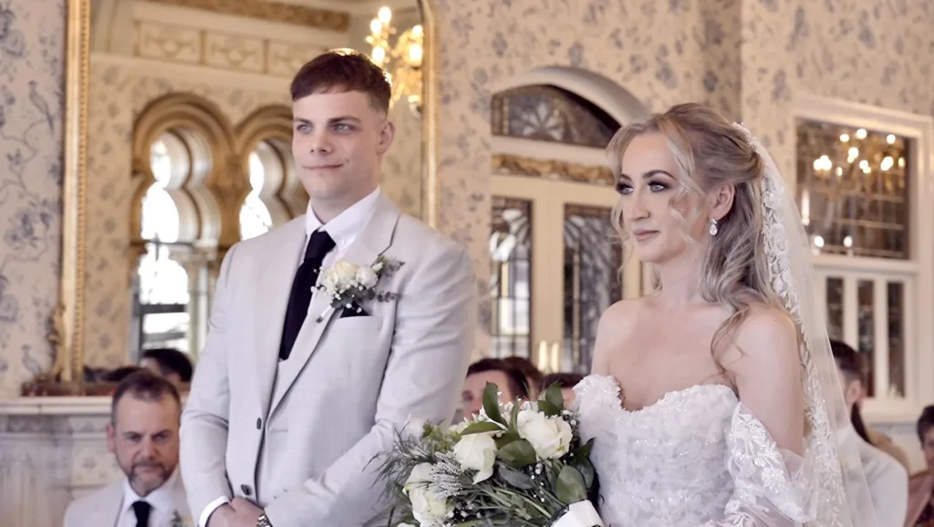 Bride and groom stand side by side at the front of an ornate ceremony room, guests behind them