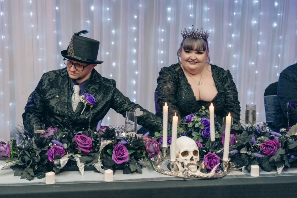 Bride and groom in black at the top table, behind purple roses and tall white candles