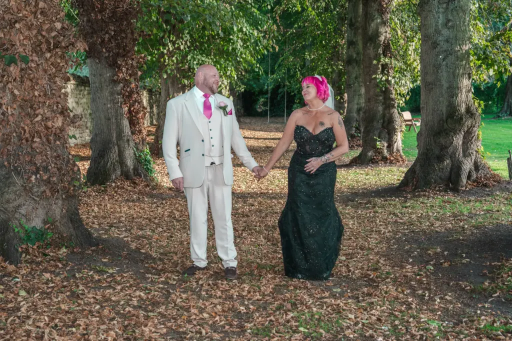 Bride and groom hold hands under tall trees on a carpet of autumn leaves