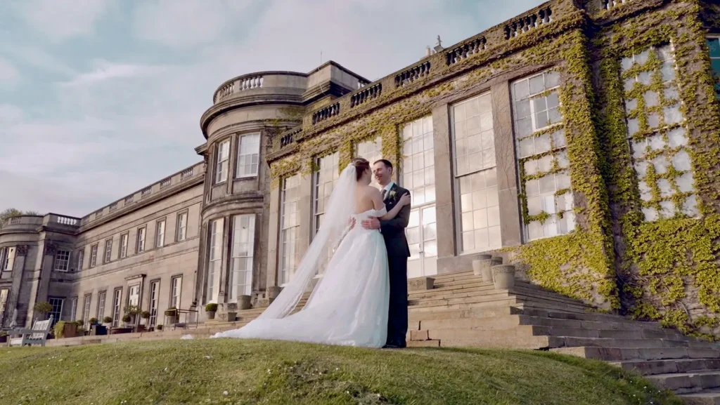 Bride and groom embrace on the lawn below a grand ivy-covered country house
