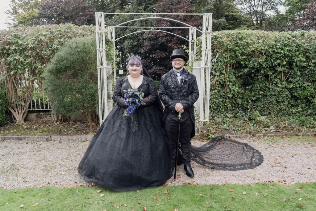 A bride in a black gown and a groom in a top hat beneath a white garden arch