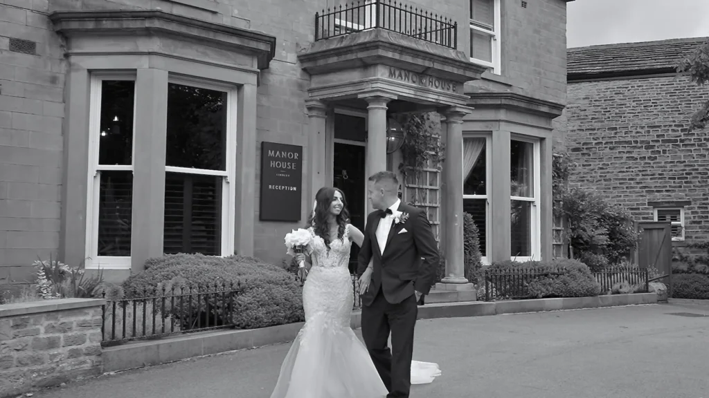 Bride and groom step out from the pillared porch of a stone manor house hotel, in black and white