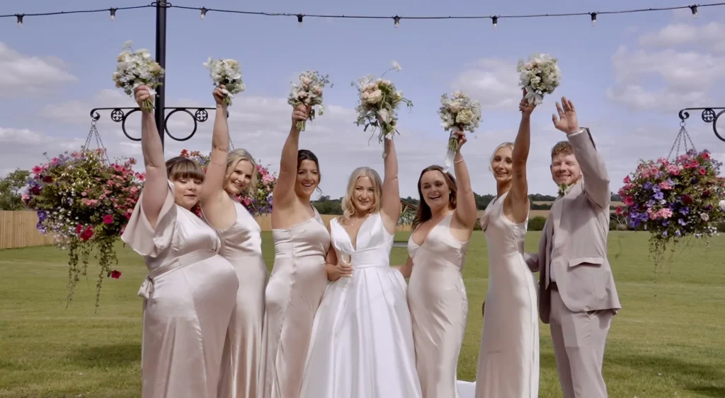 The bride and her bridal party raise their bouquets on a lawn under festoon lights
