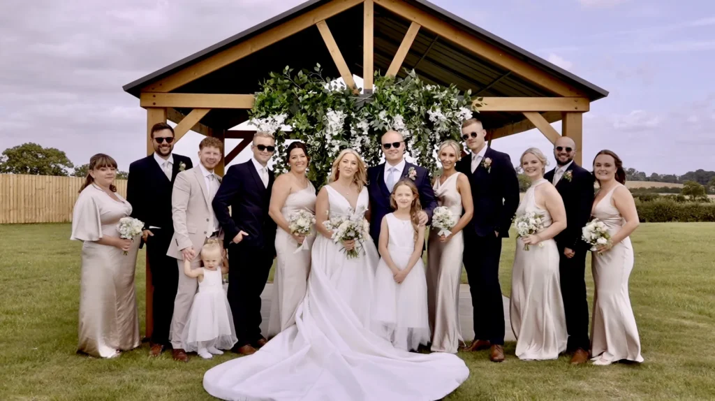 The bride and groom with their bridesmaids, groomsmen and flower girls under a wooden pavilion