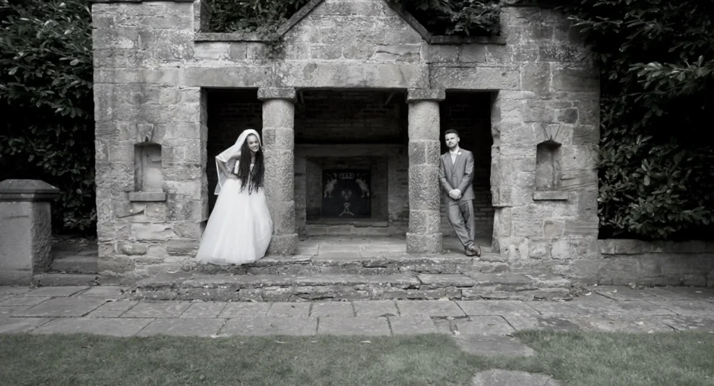 Bride and groom lean against the columns of a stone folly, one each side, in black and white