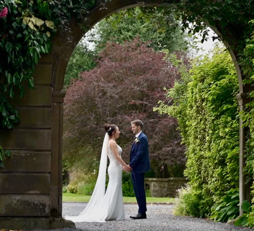 Bride and groom hold hands beneath a stone archway framed by trees