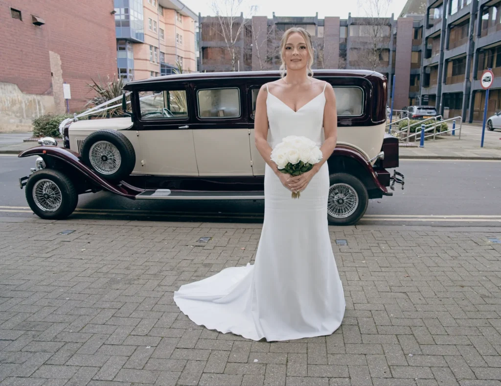 The bride holds white roses beside a burgundy and cream vintage car