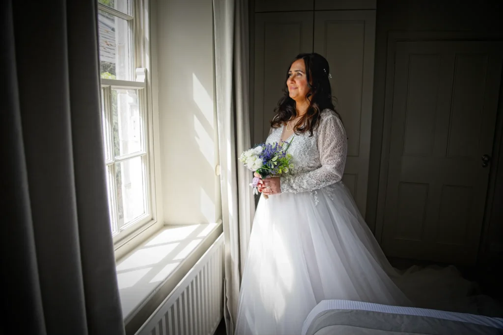 The bride holds a bouquet of white and purple flowers by a sunlit window