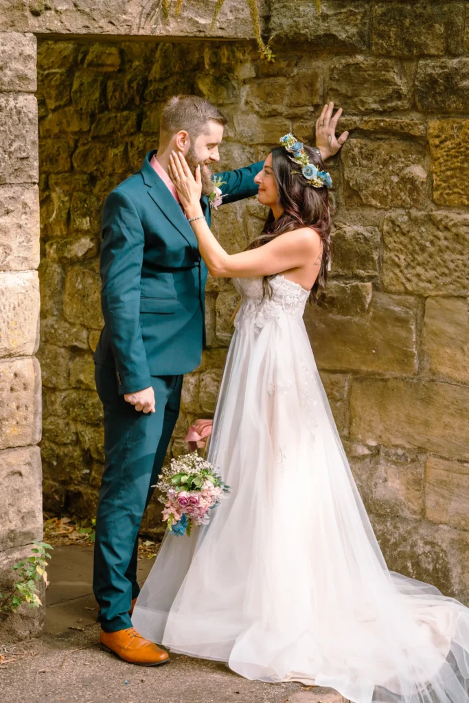 The bride cups the groom’s face as they stand against an old stone wall