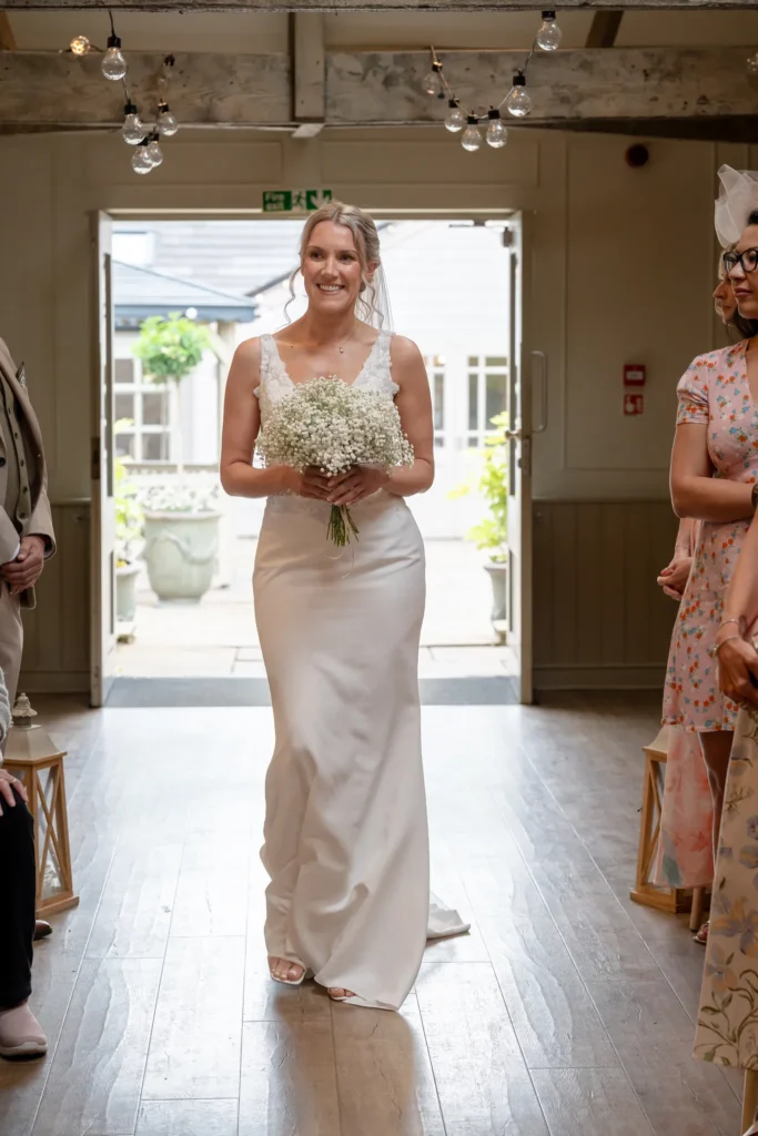 The bride smiles on her way down the aisle, festoon lights overhead