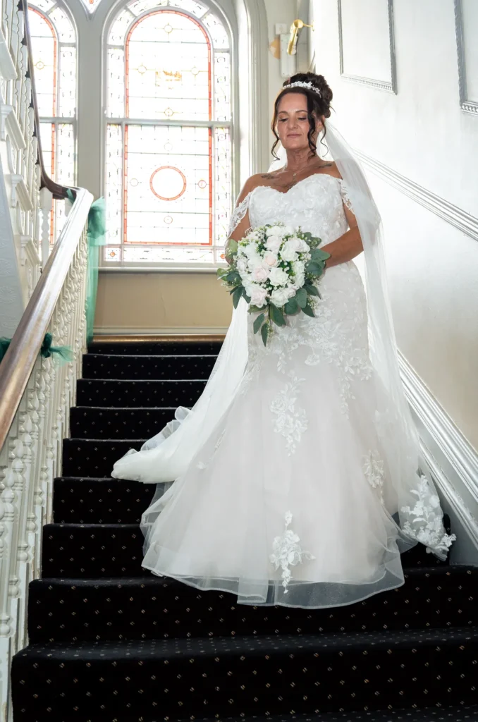 The bride walks down a carpeted staircase beneath a stained-glass window