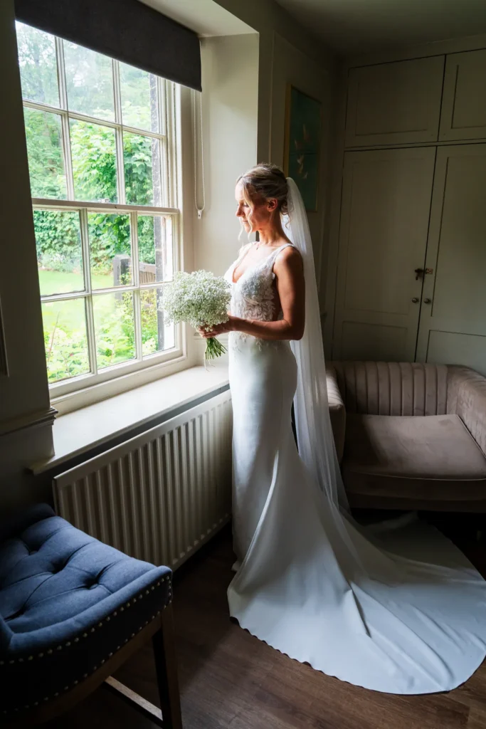 The bride holds a bouquet of gypsophila by a sash window, her veil and train on the floor