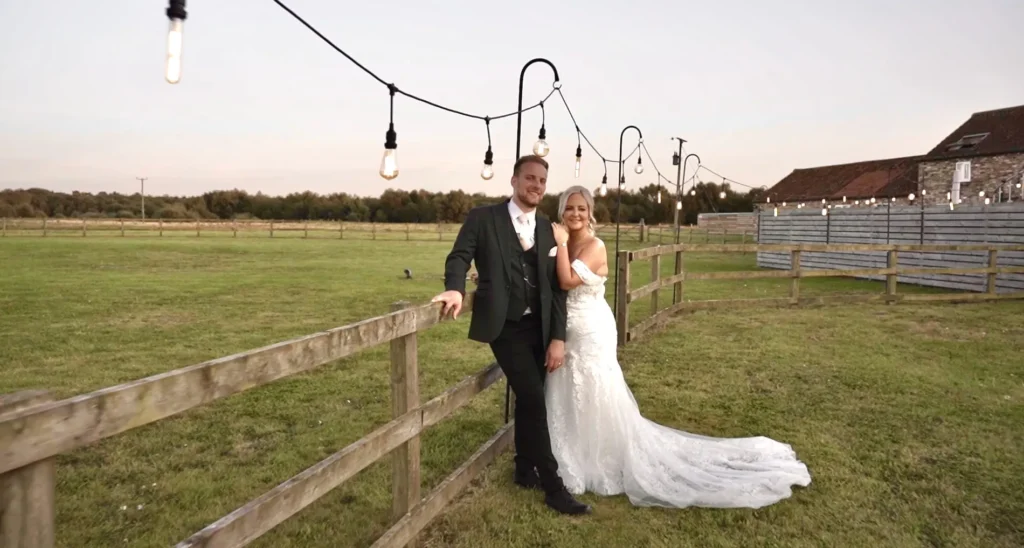 Bride and groom lean on a wooden fence under festoon lights at dusk