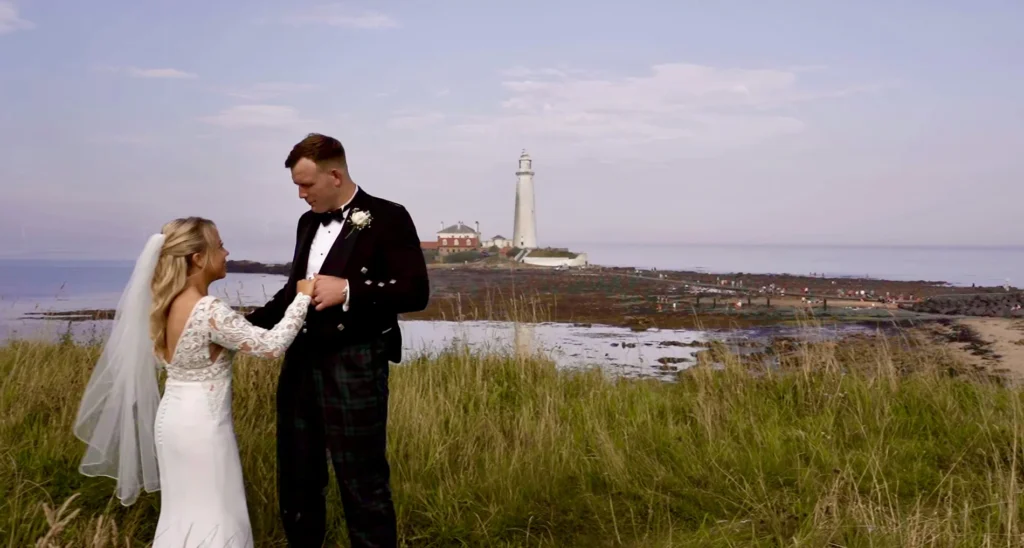 Bride and groom on a grassy clifftop, a lighthouse across the rocks behind them