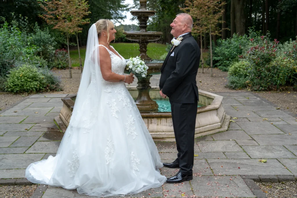 Bride and groom smile at each other beside a tiered stone fountain in a garden