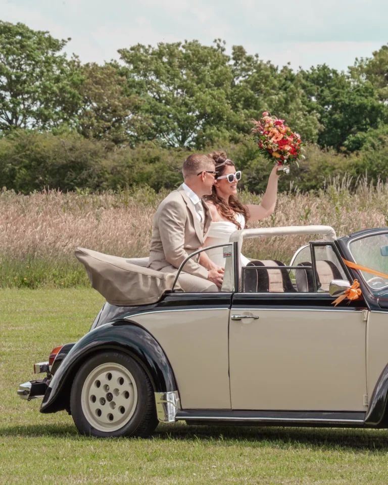 Bride and groom in sunglasses in a black and cream convertible Beetle, her bouquet raised high
