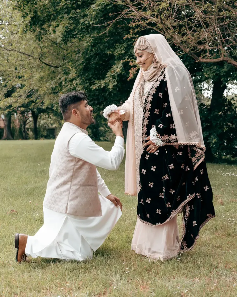 The groom kneels on the lawn to take the bride’s hand at an Asian wedding, her black velvet dress embroidered with gold