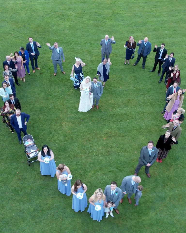 Wedding guests form a heart around the bride and groom on a lawn, seen from a drone