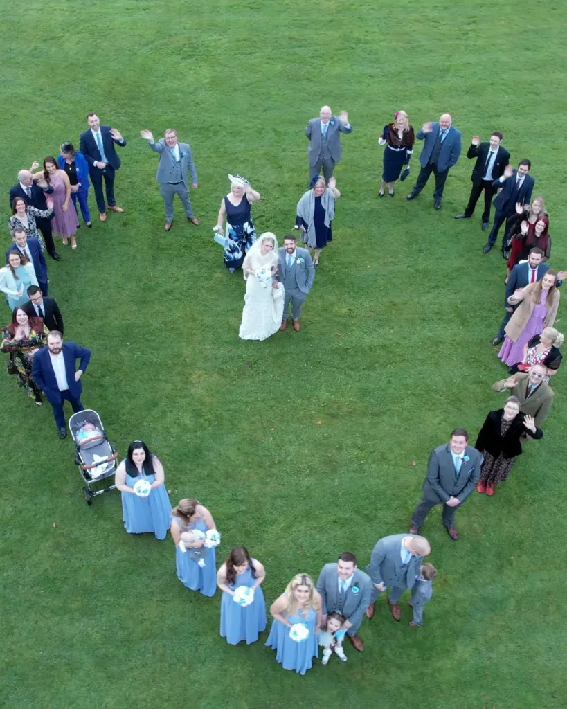 Wedding guests form a heart around the bride and groom on a lawn, seen from a drone