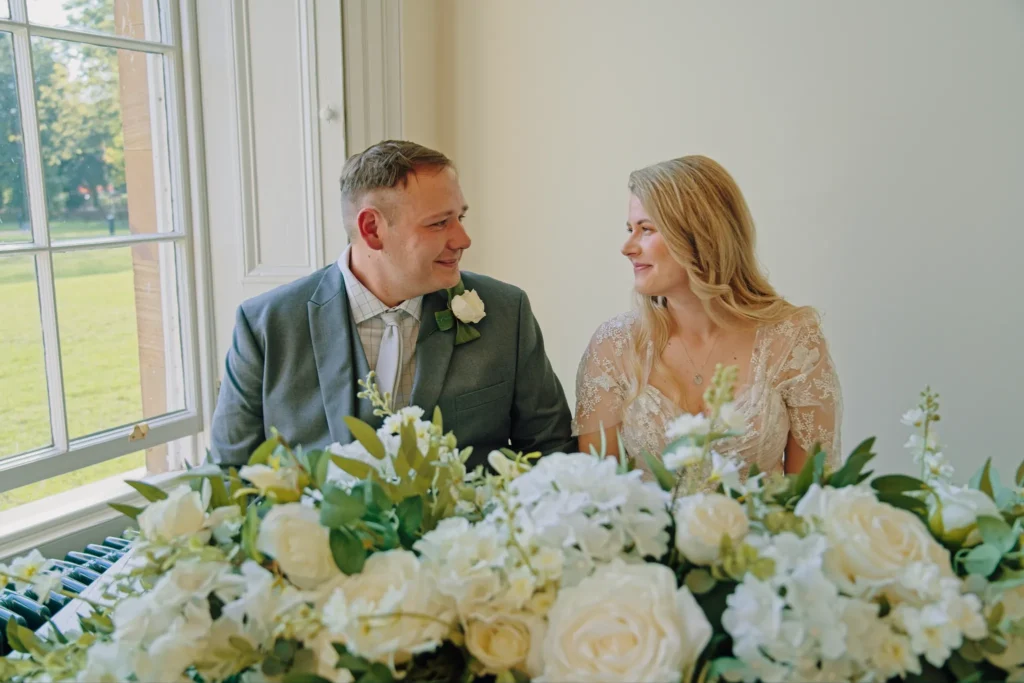 Bride and groom smile at each other behind a table of white roses by a tall window
