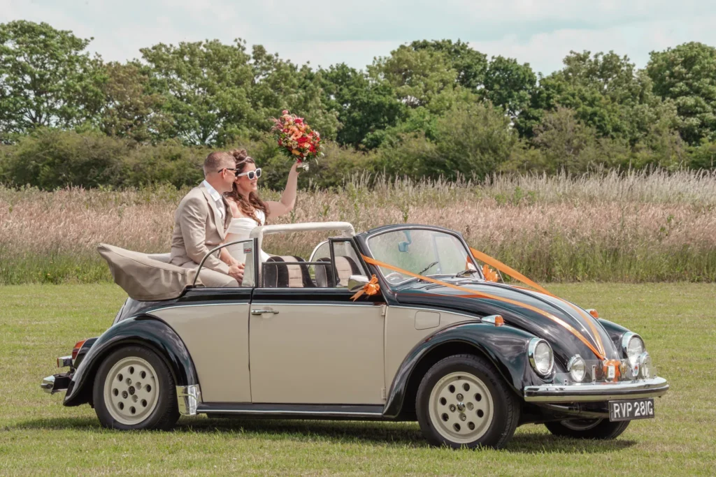 Bride and groom in sunglasses wave from a black and cream convertible Beetle