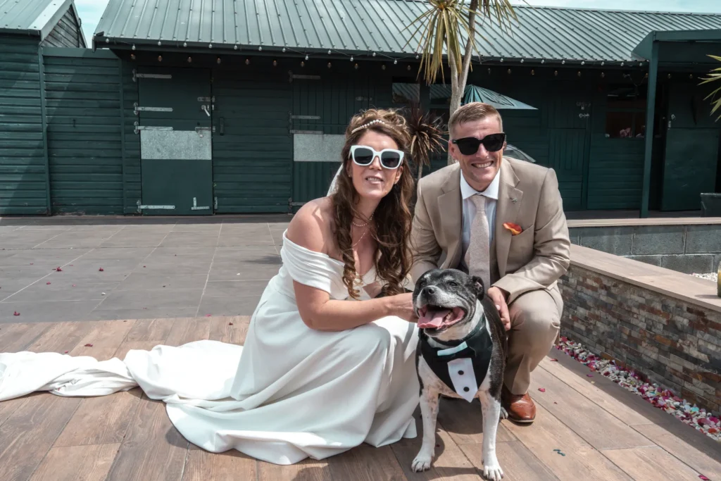 Bride and groom in sunglasses crouch beside their dog in a bow tie