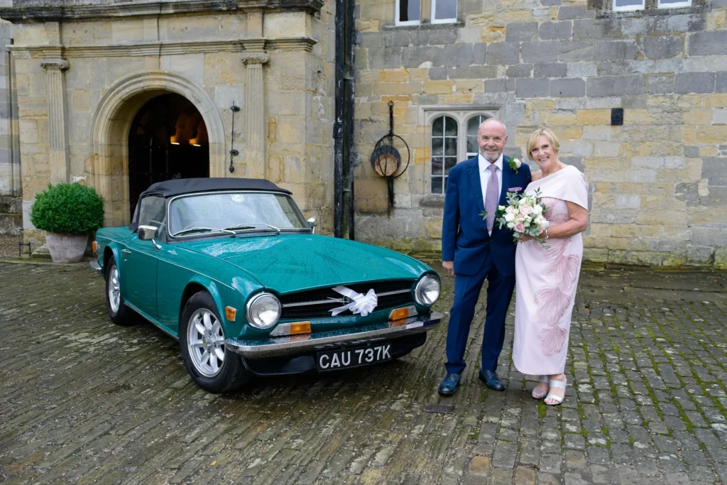 Bride and groom beside a green vintage sports car in the cobbled courtyard of a stone manor