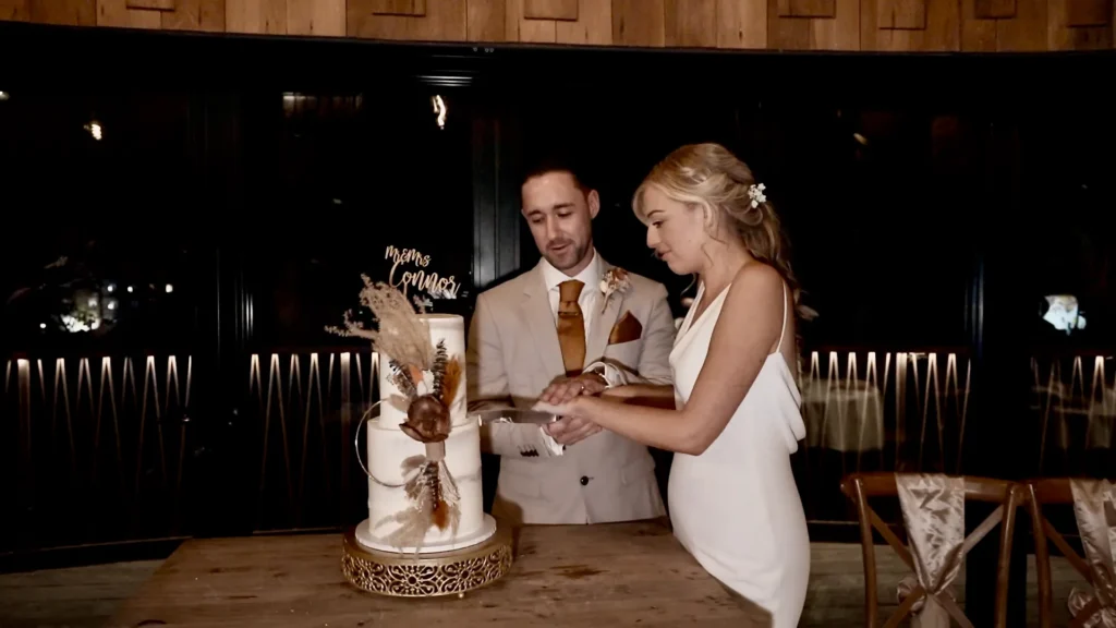 Bride and groom cut a tall white wedding cake dressed with dried grasses, in a dark wood-panelled room