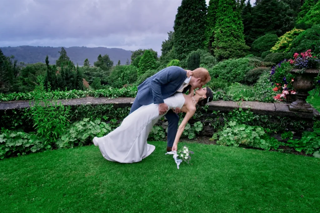 The groom dips the bride for a kiss on a garden lawn, hills behind