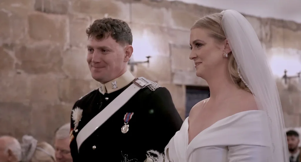 A bride in an off-the-shoulder gown and long veil and a groom in military dress uniform stand together at their ceremony, an old stone wall and their guests behind them