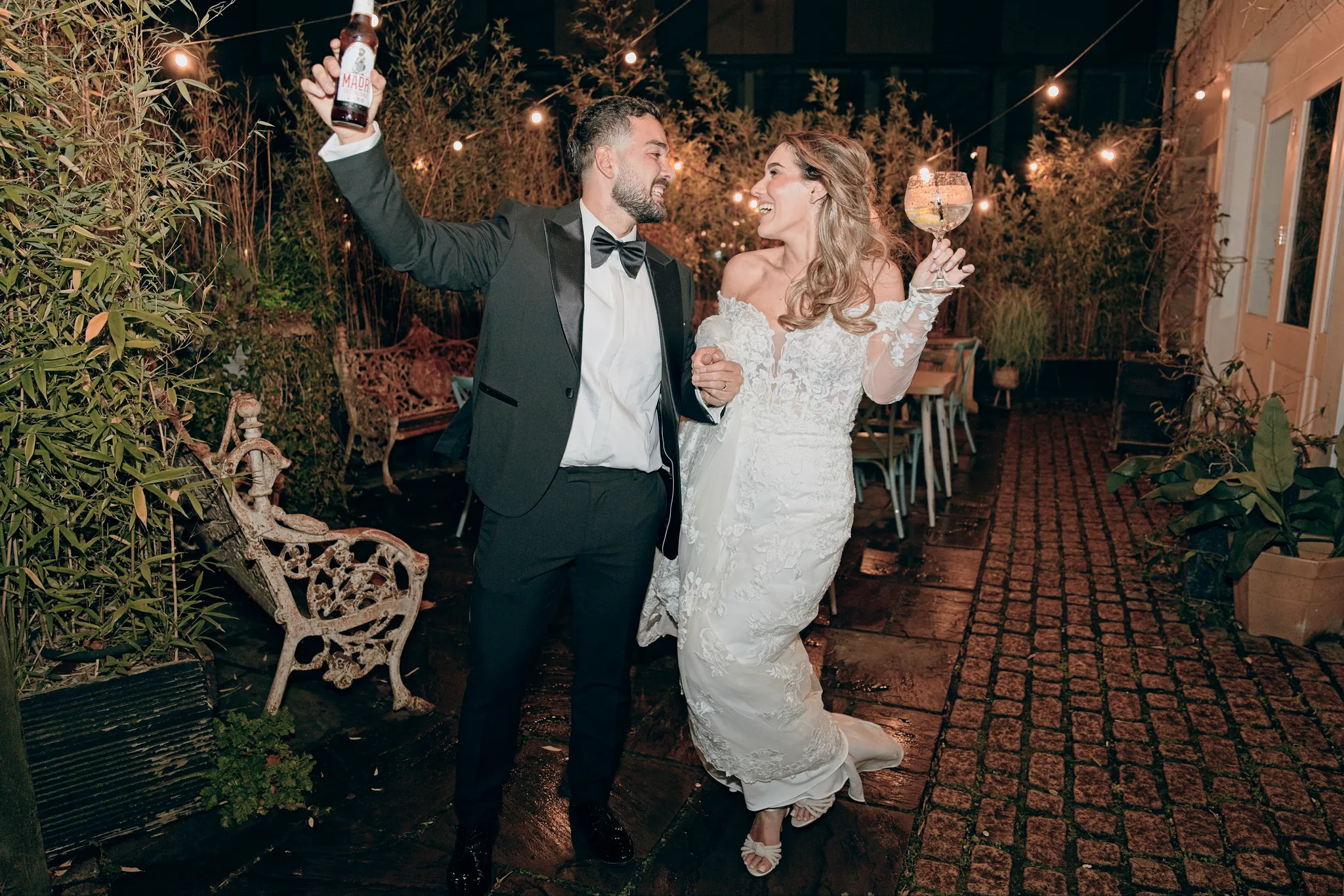 Bride and groom laugh and raise their drinks in a courtyard strung with festoon lights at night