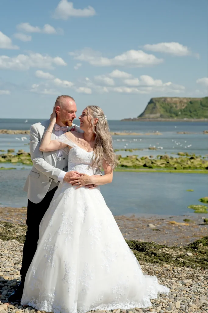 Bride and groom embrace on a pebble beach, a headland across the bay