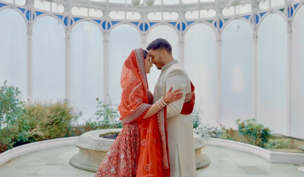 Bride and groom touch foreheads in a domed white conservatory at an Asian wedding, her red veil edged in gold