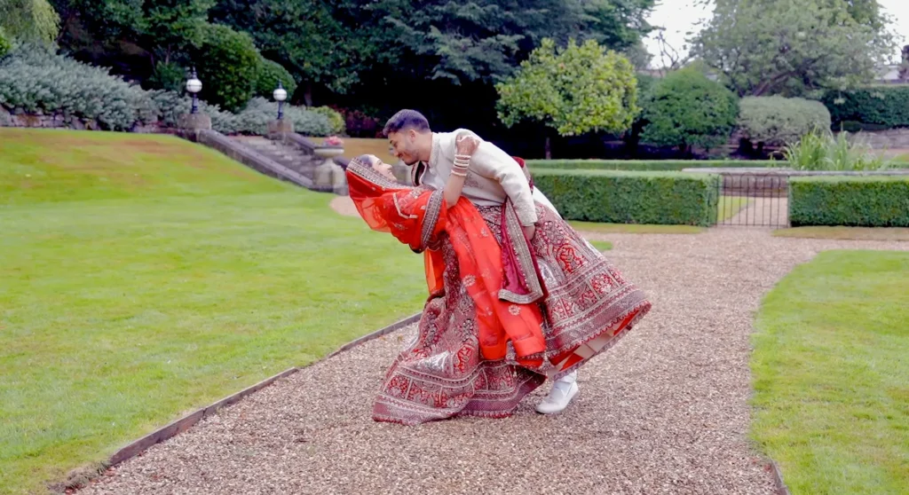 At an Asian wedding, the groom in a cream sherwani dips the bride in a red and gold lehenga on a gravel path in formal gardens