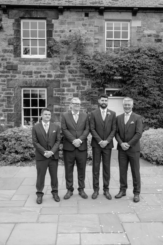 The groom and his groomsmen outside a stone house, in black and white
