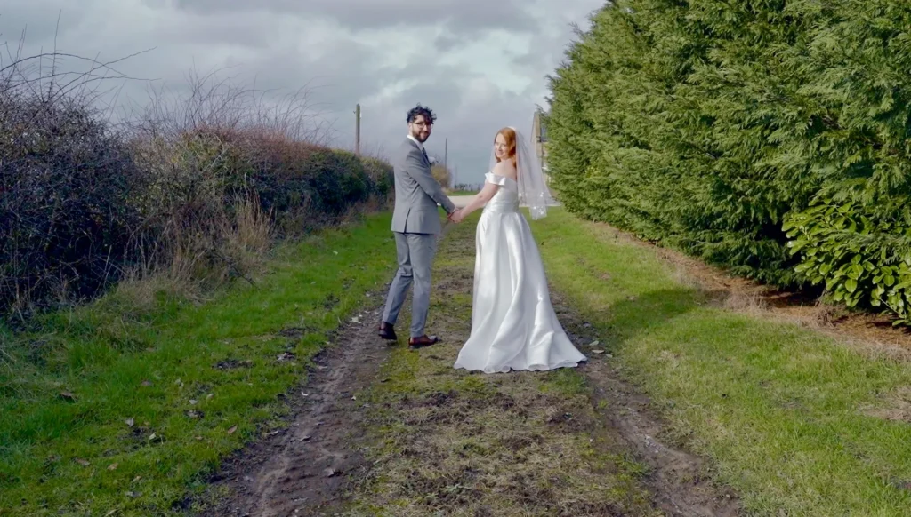 Bride and groom hand in hand on a grassy farm track between tall hedges