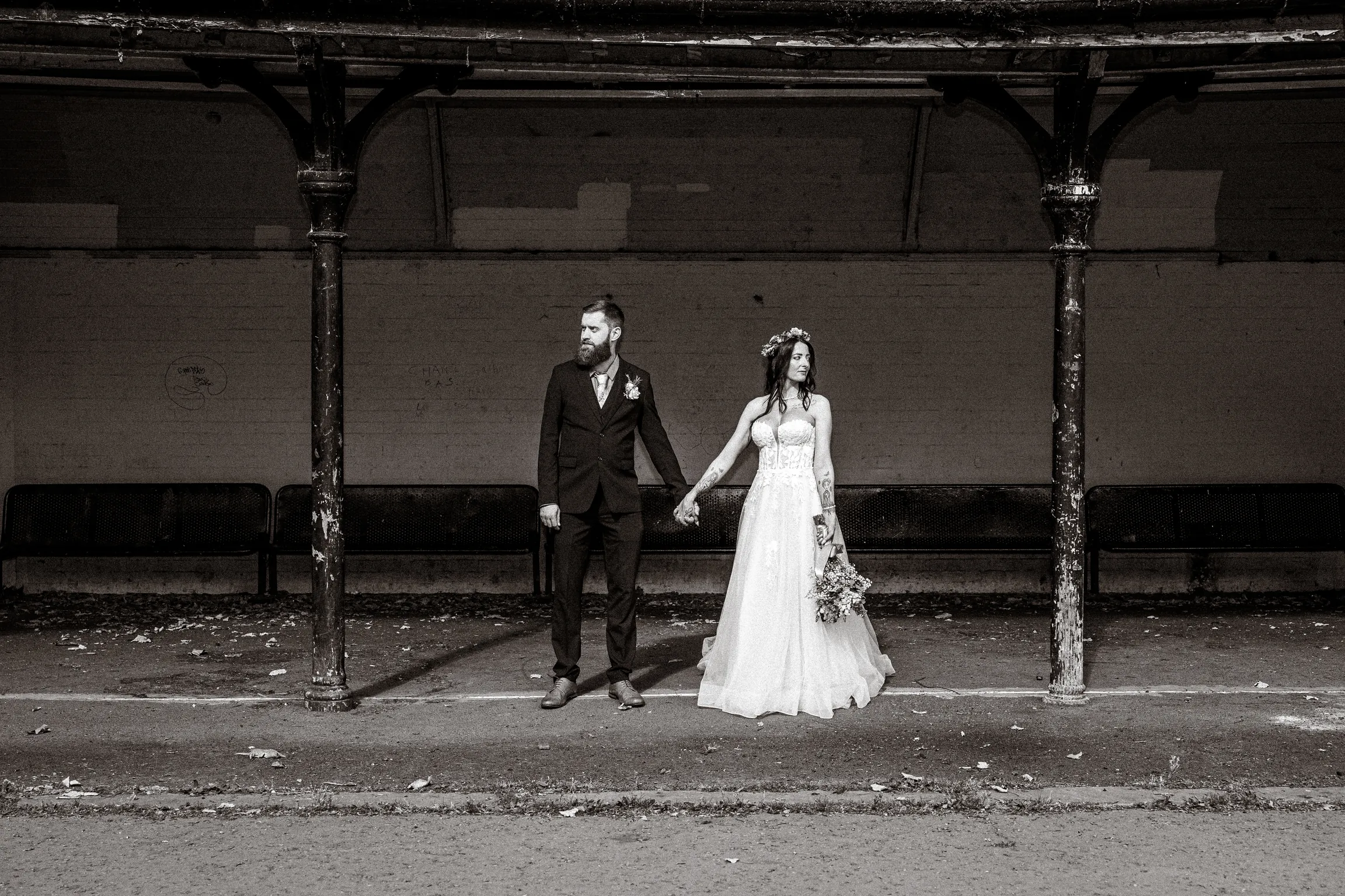 Bride and groom hold hands under an old iron shelter, looking in opposite directions, in black and white