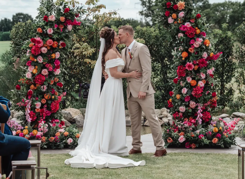 Bride and groom kiss between pillars of bright flowers at an outdoor ceremony