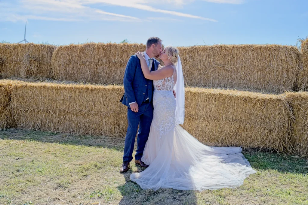 Bride and groom kiss in front of stacked straw bales under a blue sky