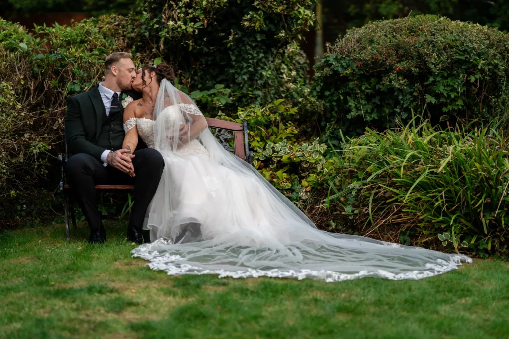 Bride and groom kiss on a garden bench, her long veil spread across the lawn