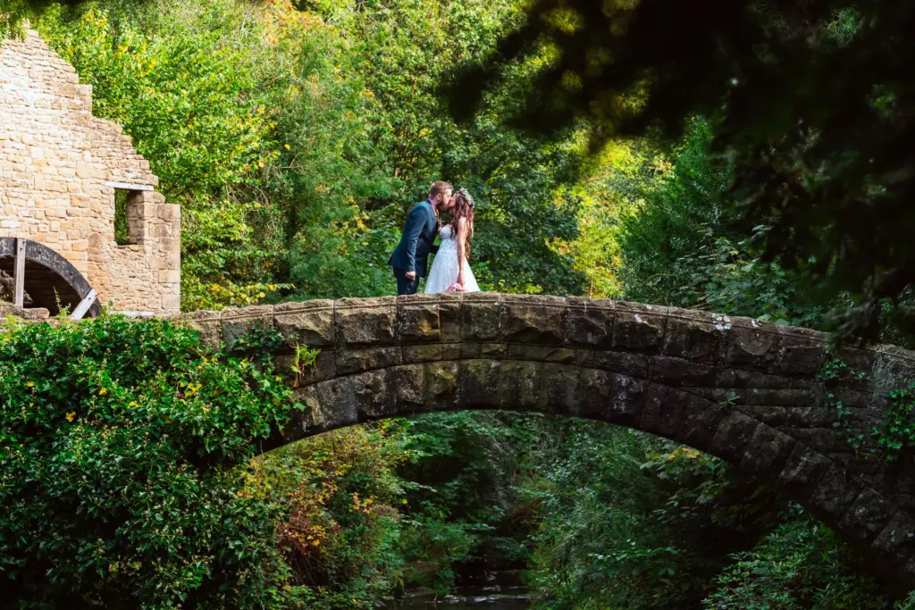 Bride and groom kiss on an old stone bridge beside a ruined mill and its water wheel
