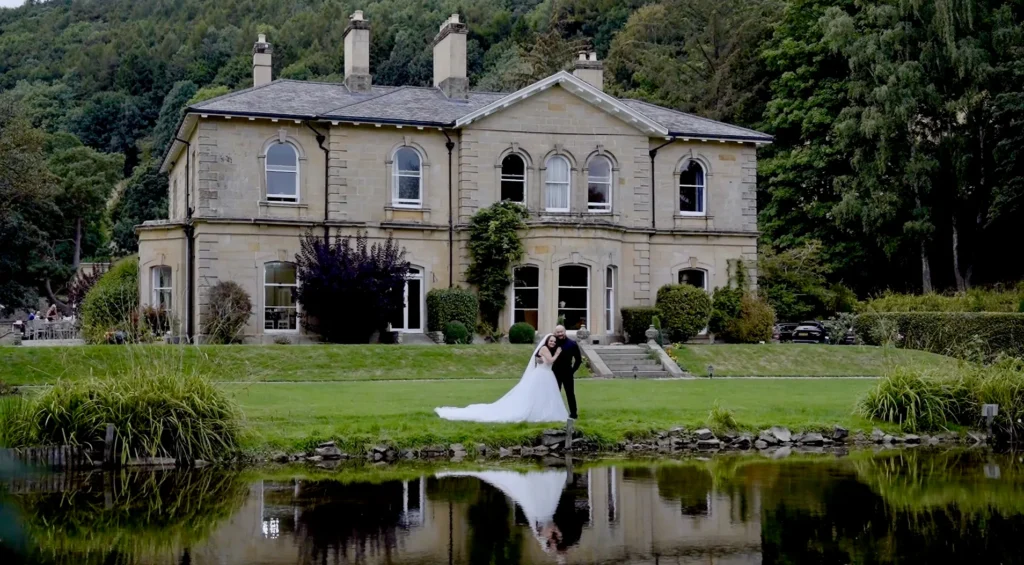 Bride and groom on a lawn by a lake in front of a stone country house, reflected in the water