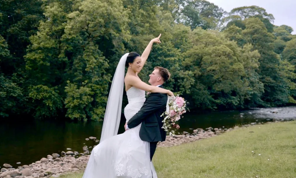 The groom lifts the bride beside a river, her arm raised to the sky