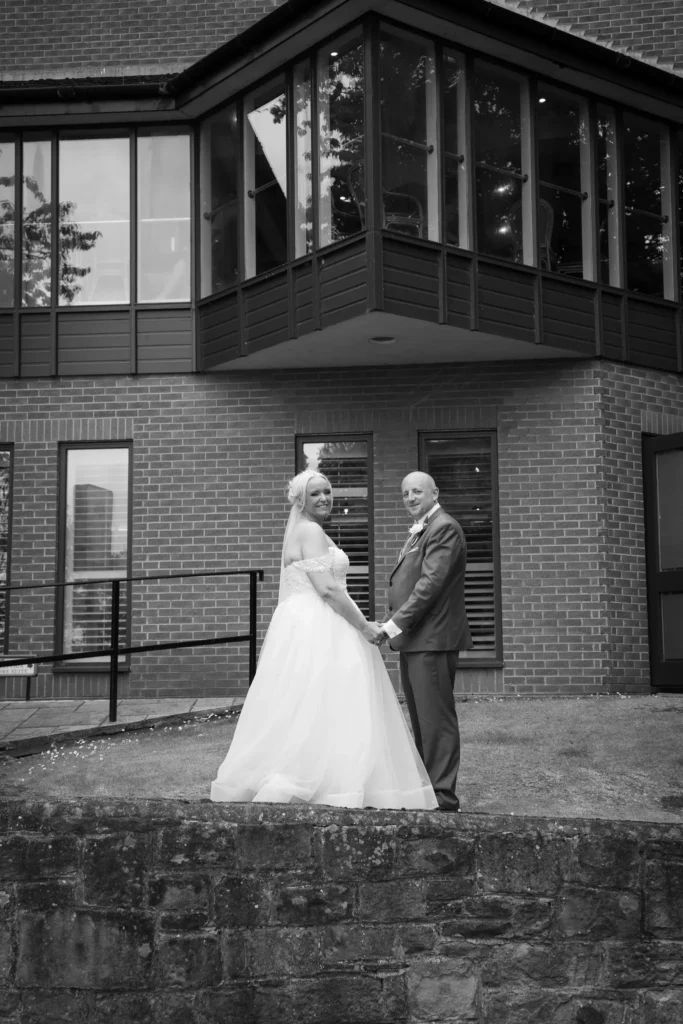 Bride and groom hand in hand look back over their shoulders outside a brick building, in black and white