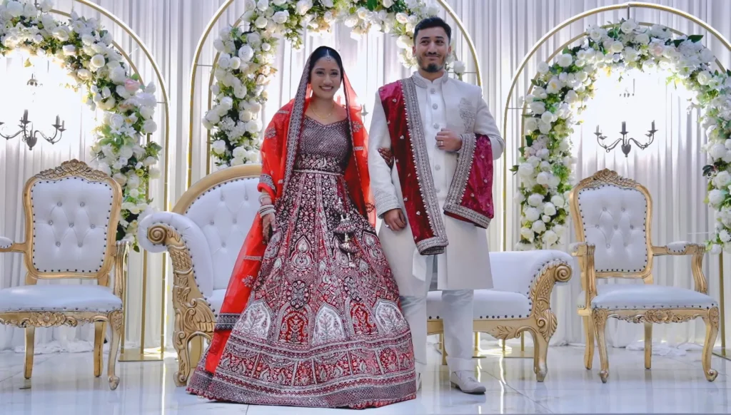 At an Asian wedding, the bride in a red lehenga and the groom in a white sherwani stand on a stage between arches of white flowers