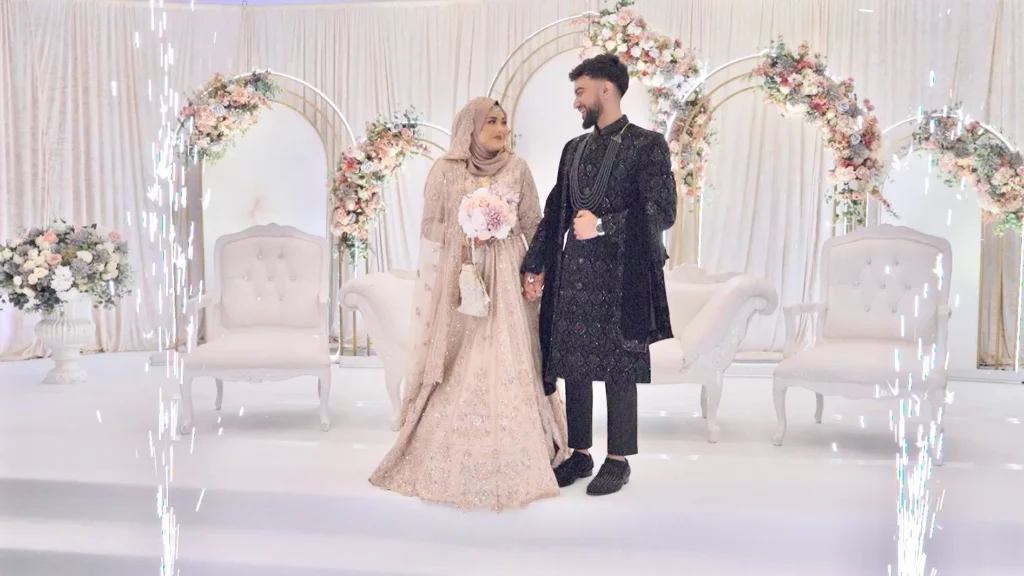 Bride and groom hold hands on a white stage with floral arches and sparkler fountains at an Asian wedding
