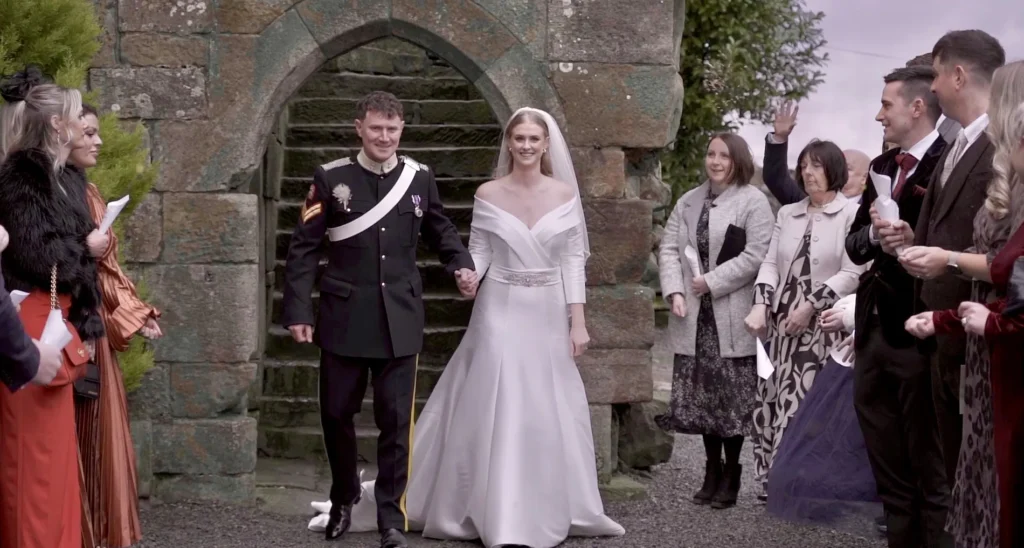 Bride and groom walk out through an old stone archway as their guests cheer